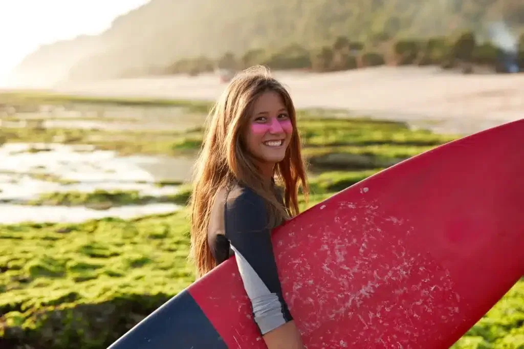 Mujer sonriente con tabla de surf roja en la playa, con maquillaje facial rosa, rodeada de naturaleza y luz dorada del atardecer.