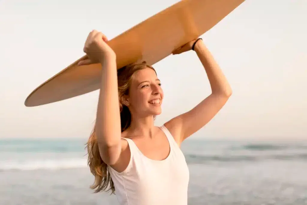 Mujer sonriente sosteniendo una tabla de surf sobre su cabeza en la playa, con el mar y el cielo de fondo.
