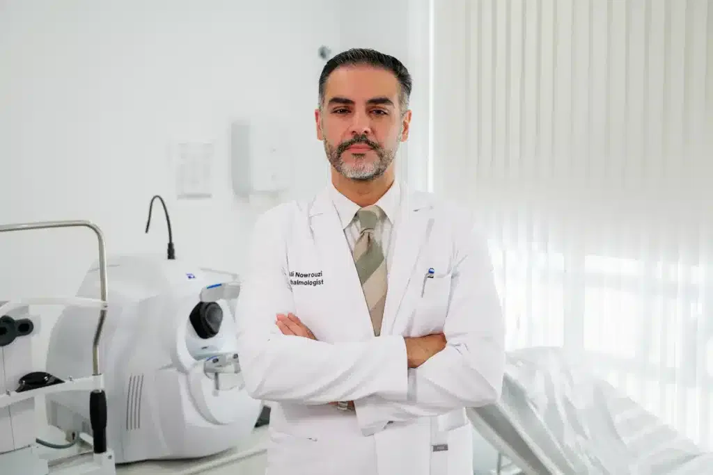 Médico en un consultorio dental, vestido con bata blanca, de brazos cruzados, con equipo dental en el fondo.