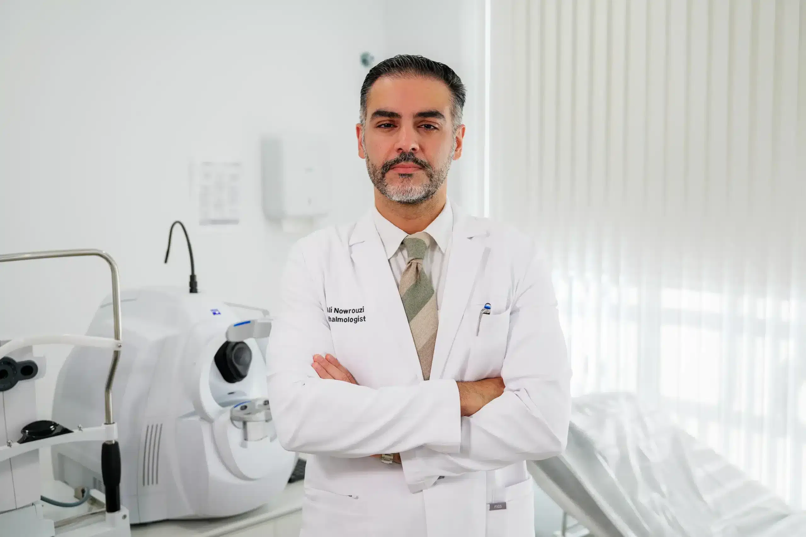Médico en un consultorio dental, vestido con bata blanca, de brazos cruzados, con equipo dental en el fondo.
