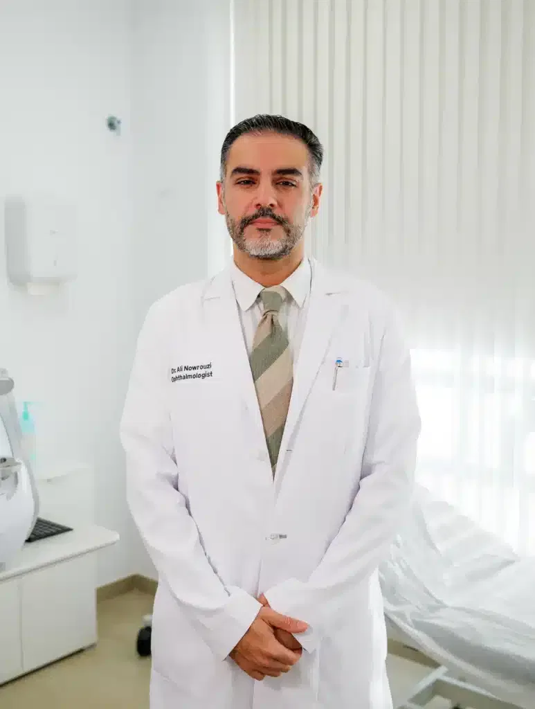 Médico de pie en consultorio, vestido con bata blanca y corbata, mostrando profesionalismo en un ambiente clínico.