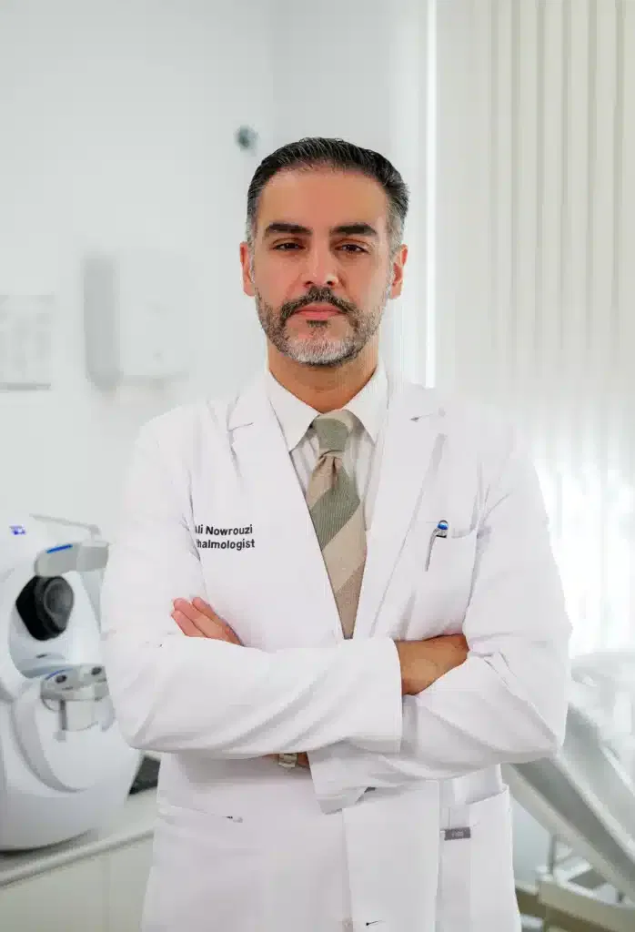 Médico de pie con bata blanca, brazos cruzados, en un consultorio moderno con equipo médico al fondo.