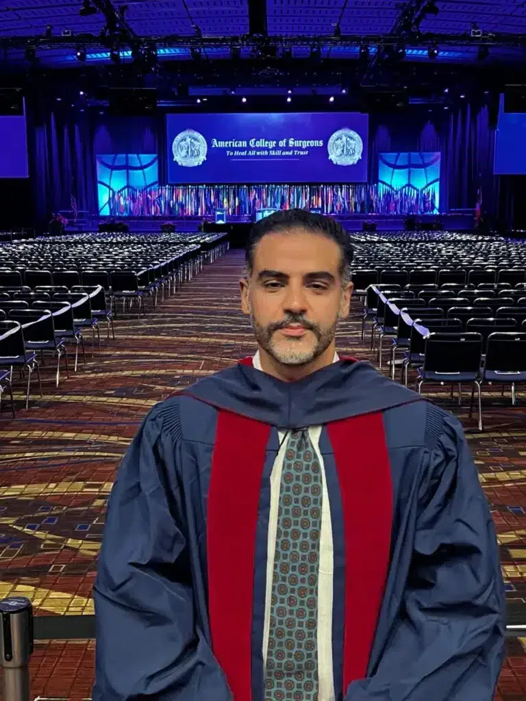 Hombre graduado con toga y birrete posando frente al escenario de una ceremonia de graduación en el Southern College of Optometry. Fondo de asientos vacíos y decoraciones del evento.