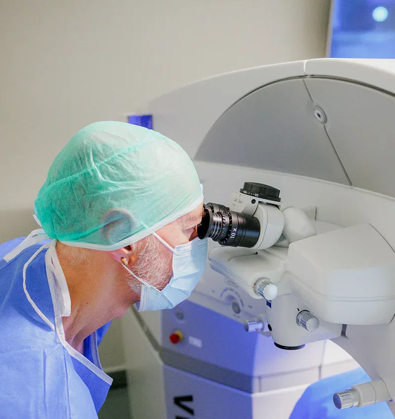 Médico utilizando un microscopio en un entorno clínico, con gorro y mascarilla, enfocado en un procedimiento quirúrgico.