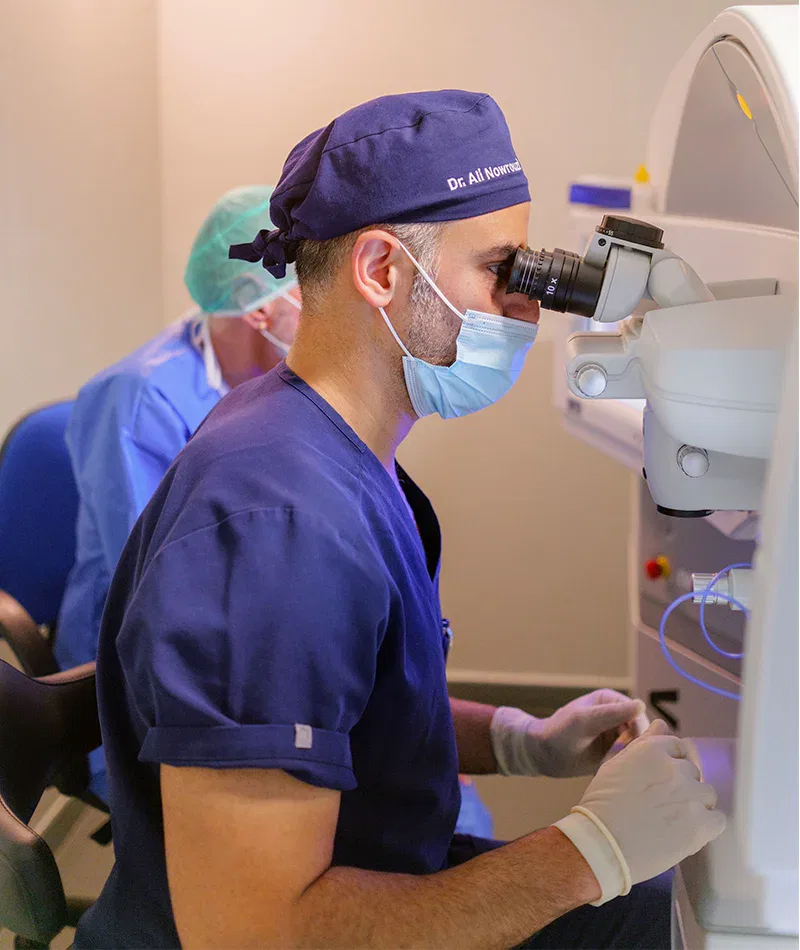 Médico utilizando un microscopio en un entorno clínico, enfocado en procedimientos oftalmológicos, con equipo de protección y guantes.