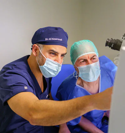 Oftalmólogo en Marbella 14 El Dr. Ali Nowrouzi y un compañero, uno con gorra y bata azul y otro con gorra verde, observan atentamente una pantalla durante una consulta médica, ambos usando mascarillas.